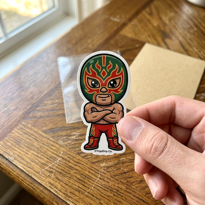 Sticker of a mexican luchador character with a green mask and red outfit held by a hand on a wooden surface.