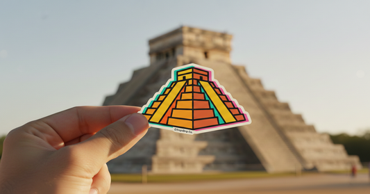 A hand holds a vibrant vinyl sticker of a stylized Aztec pyramid in the foreground, with the massive stone Chichén Itzá temple blurred in the background under soft golden hour sunlight. The sticker features bright orange, yellow, and teal geometric patterns with a thick white border and the '@TropiDrip Co.' logo.