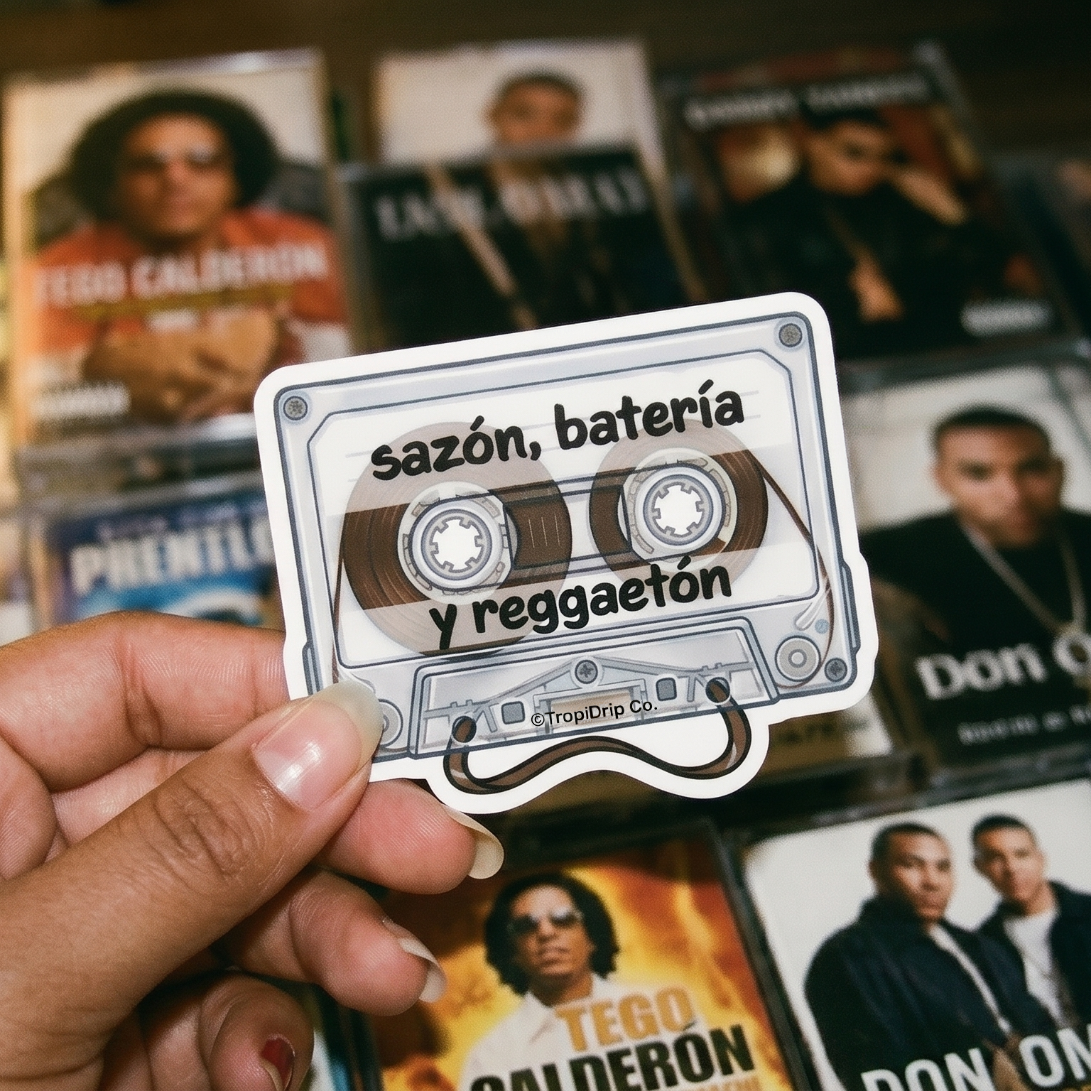 Female hand holding a cassette tape sticker with text in front of reggaeton music album covers.